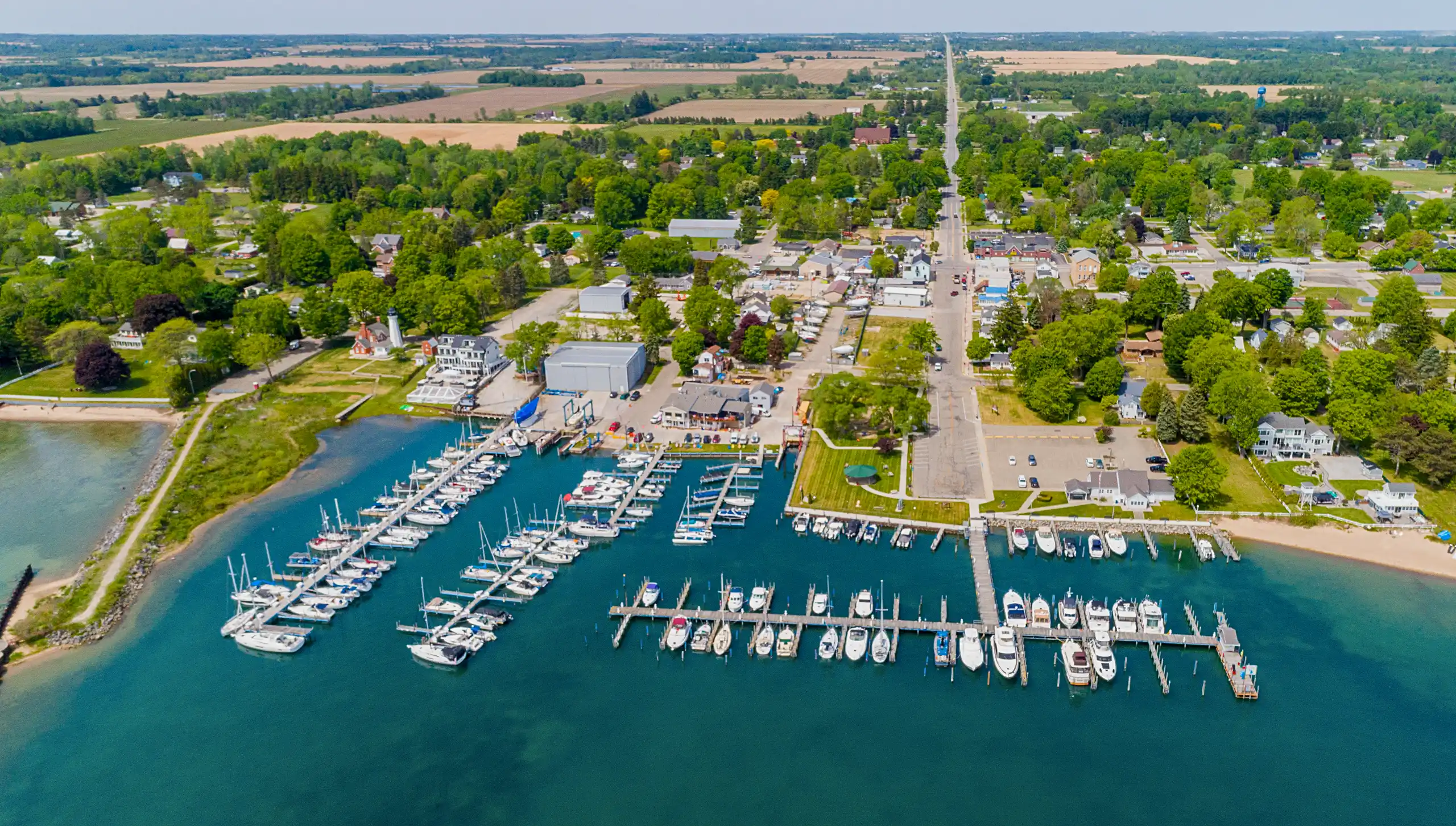 A vibrant aerial view of a coastal village. Boats are docked in the teal marina, bordered by dense green trees, leading to an inland road surrounded by farmland.