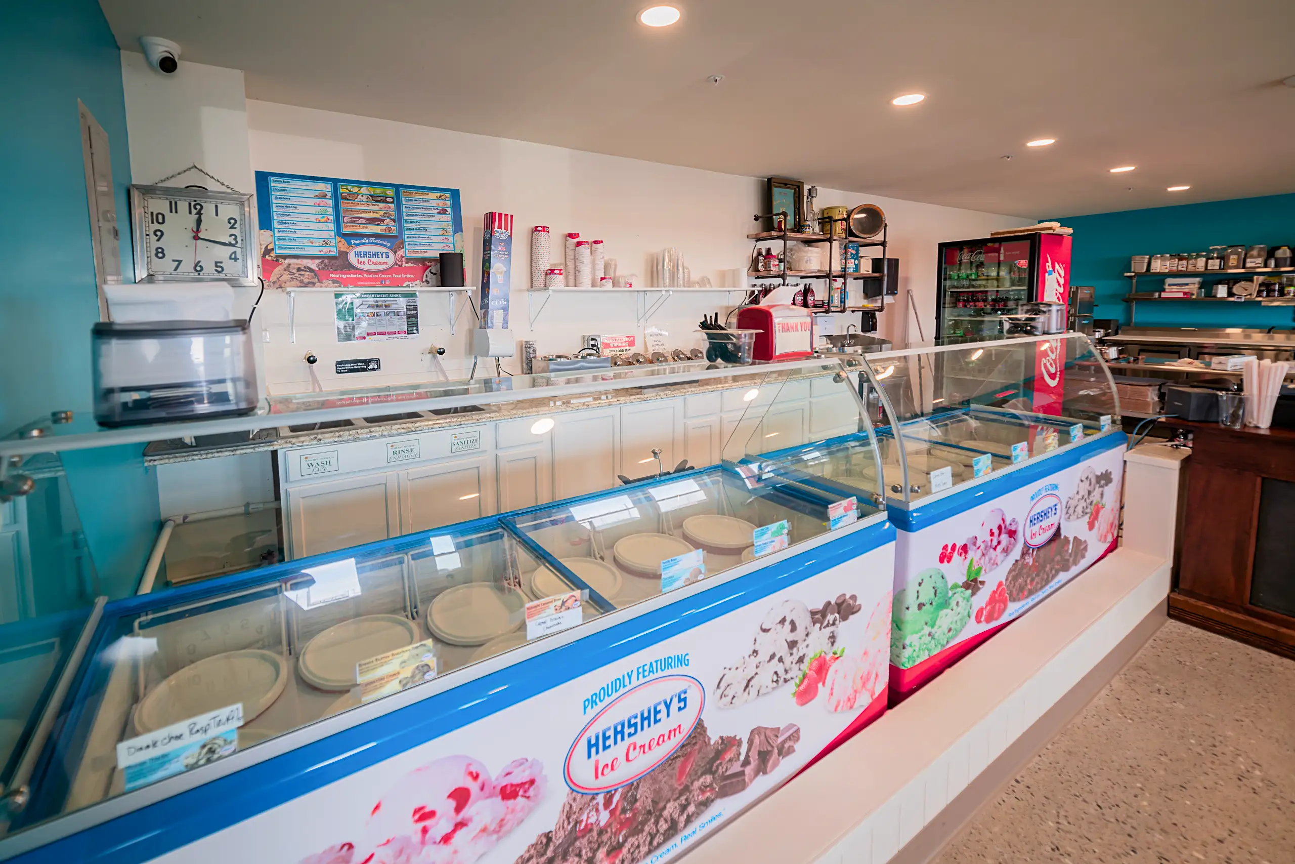A brightly lit ice cream parlor counter with a long display freezer showcasing Hershey's ice cream flavors and a clean service area with wash stations.