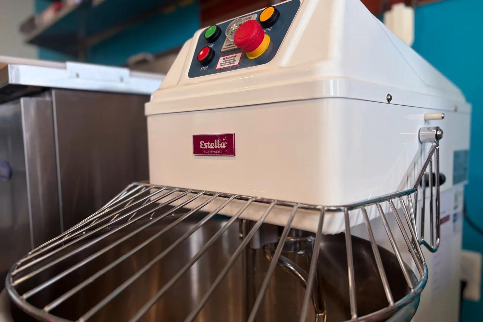 A bright white commercial dough mixer featuring a red emergency stop button and a steel wire guard over the large mixing bowl.