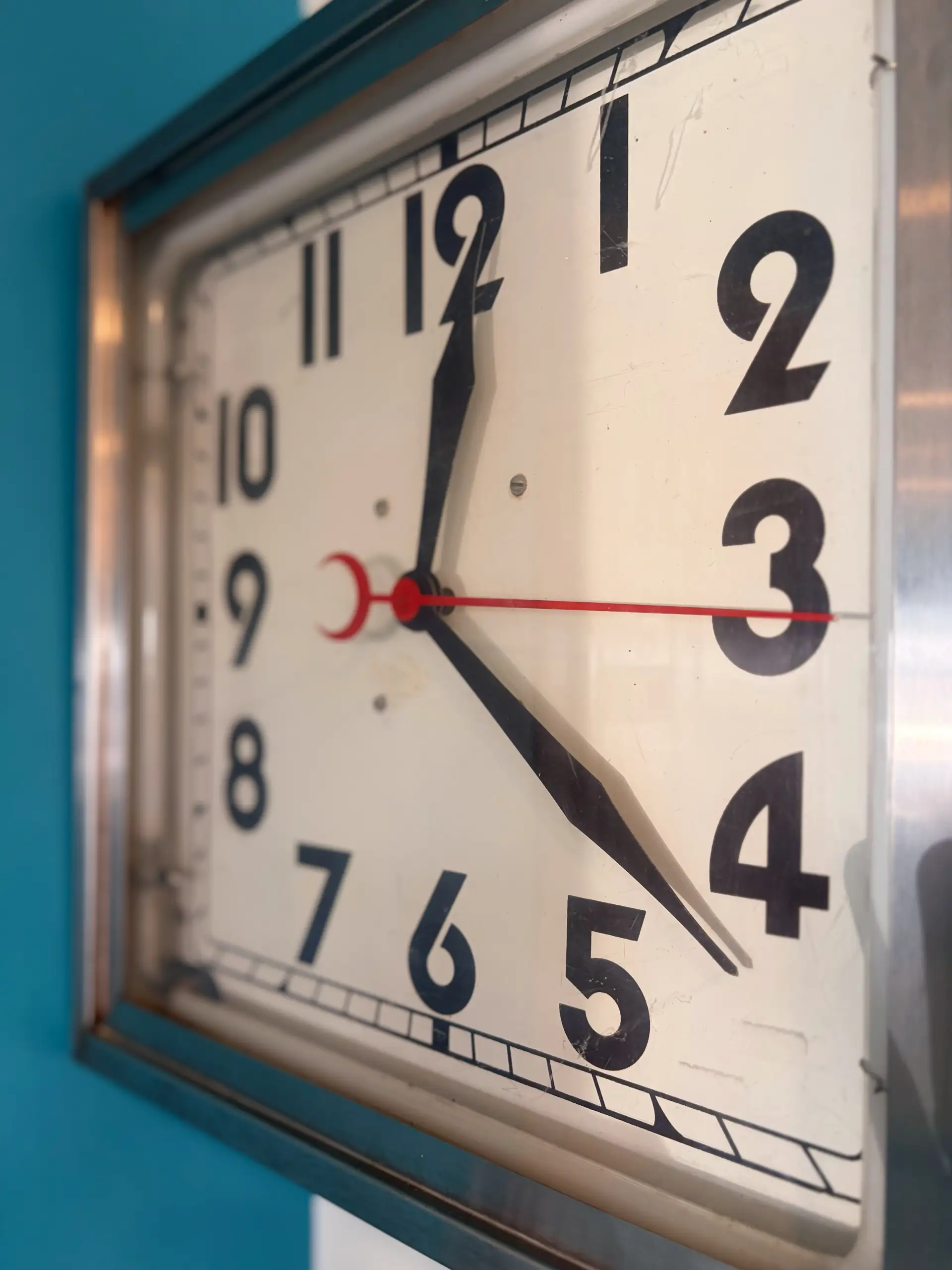 A close-up view of a vintage square wall clock with bold black numerals and a white face. The red second hand sweeps over the center, mounted on a bright turquoise wall.