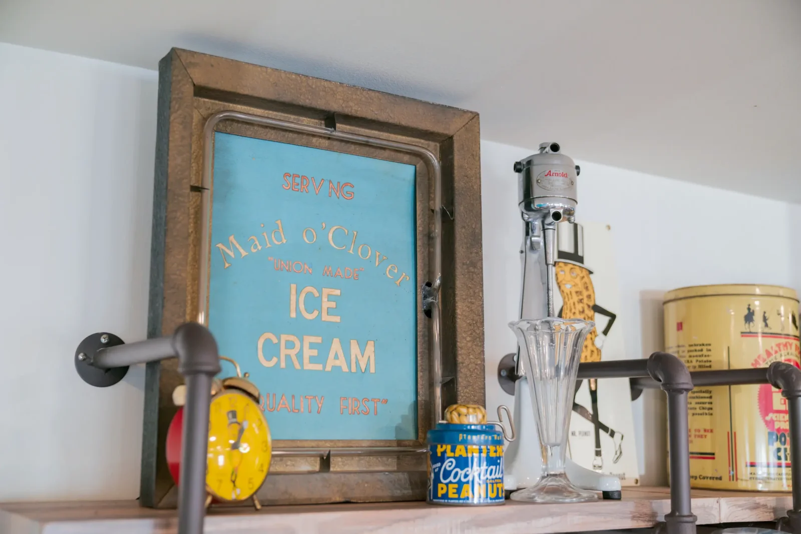 A display of vintage soda shop items featuring a Maid o'Clover ice cream sign, an Arnold milkshake mixer, and a Planters peanut tin.